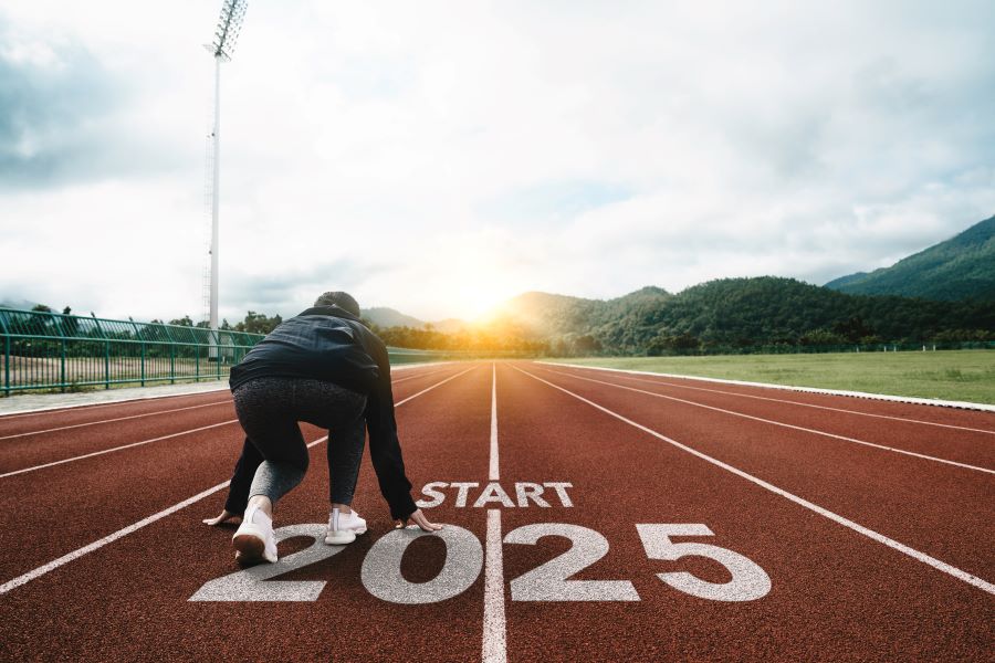 A woman in athletic gear crouches at the starting line of a running track labeled "START 2025," with mountains and a rising sun in the background, symbolizing a fresh start and new goals for the year 2025.